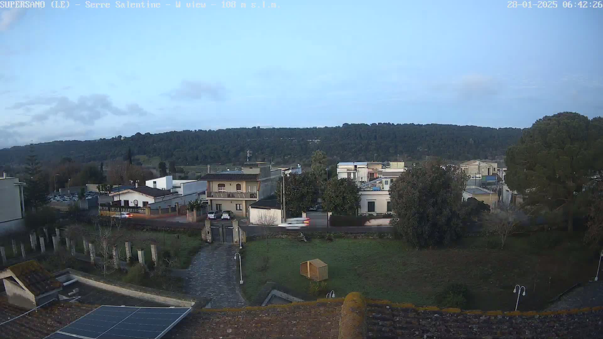 A mostly clear, dusky sky overlooks a small town nestled beside a dark green hill, with several cars visible on a road below.