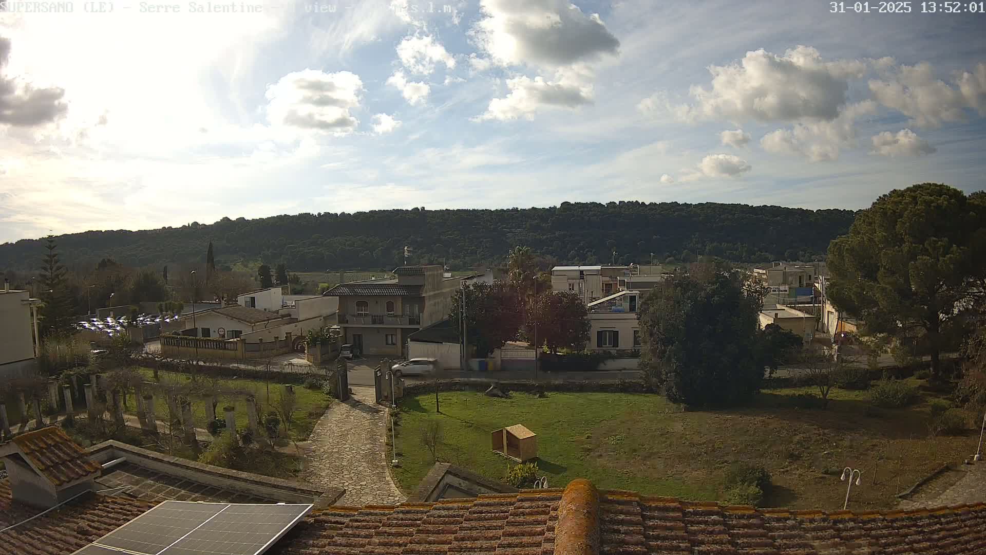 A partly sunny day reveals a hilly landscape of green trees and a residential area with houses and a paved road, viewed from a rooftop with a solar panel.