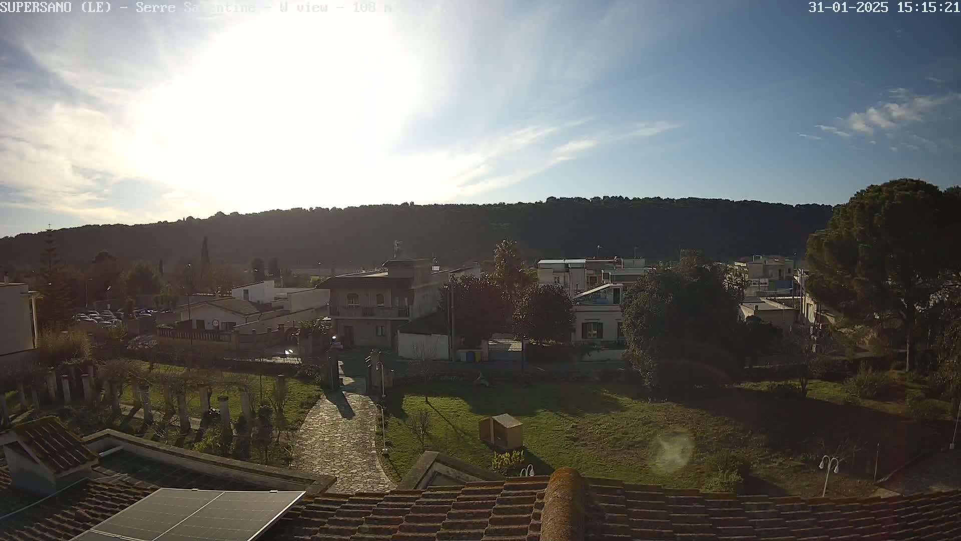 A mostly sunny day reveals a view of a small town nestled against a hillside, seen from a rooftop with solar panels.