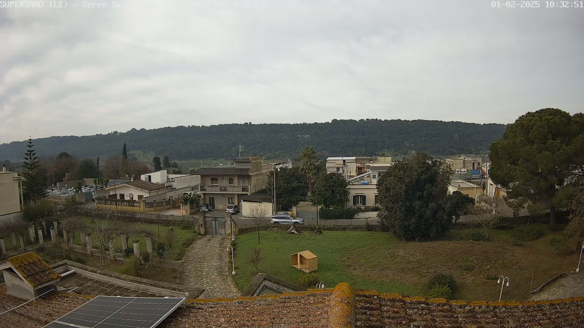 An overcast day reveals a residential area nestled against a verdant hillside, viewed from a rooftop with a solar panel.