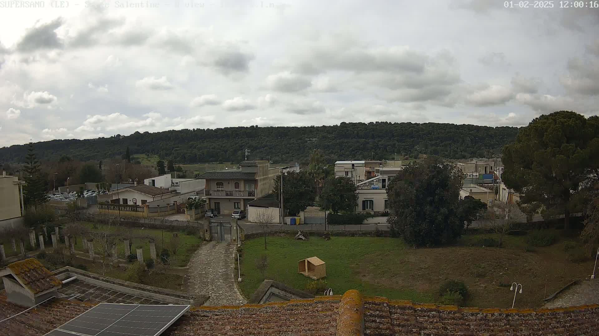 A cloudy day overlooks a residential area with low buildings nestled amongst green lawns and trees, a hill covered in dense forest in the background.