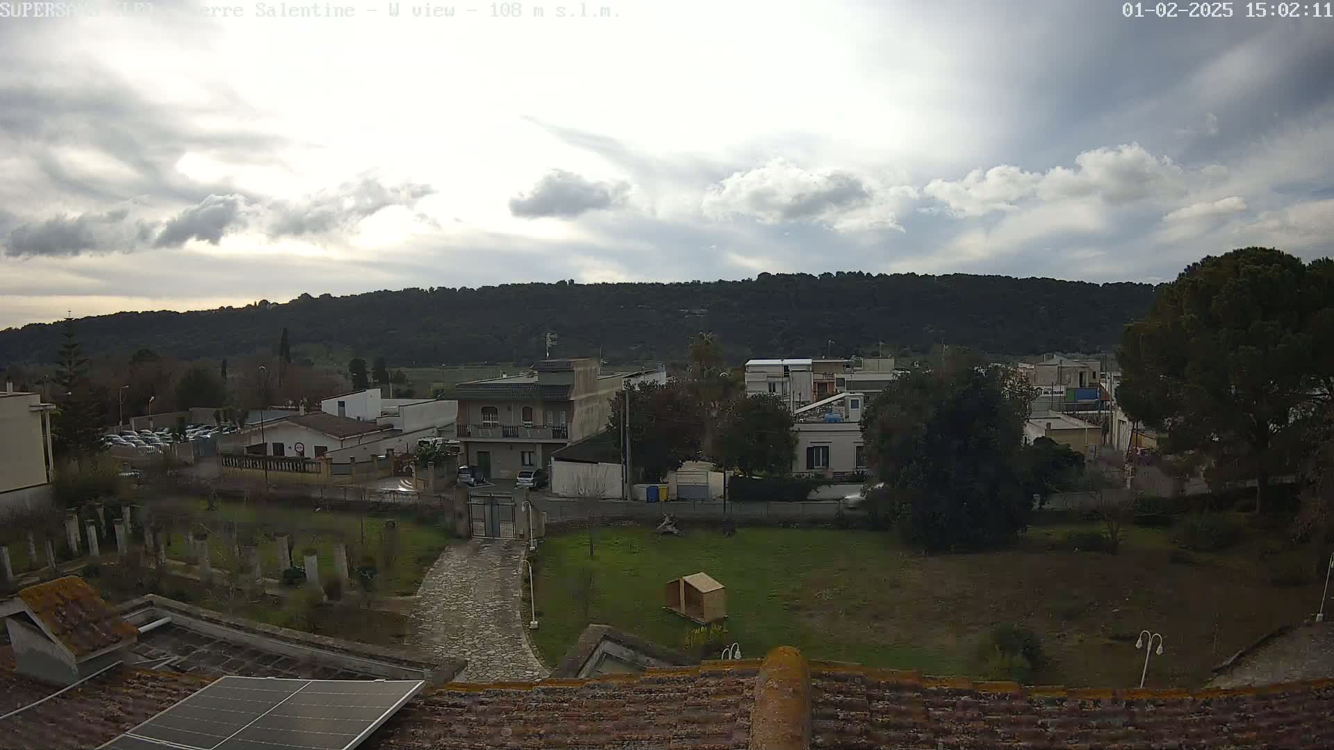 A view from a rooftop shows a partly cloudy sky over a small town nestled against a tree-covered hill.