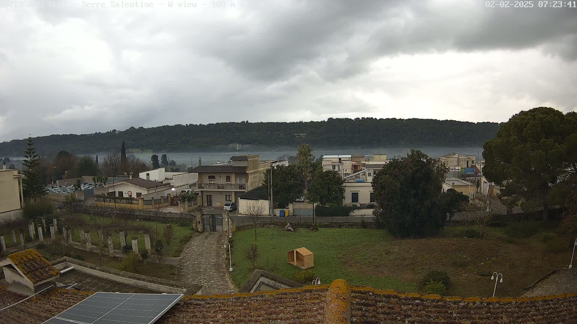 A misty, overcast day reveals a residential area nestled beside a low, wooded hillside, with a solar panel visible on a nearby roof.