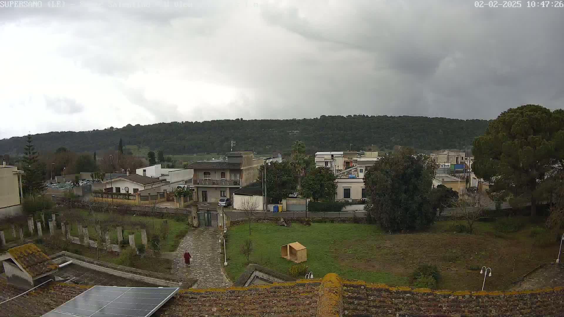 A cloudy day overlooks a residential area with houses and green spaces, a person walking on a path, and a hill covered in trees in the background.