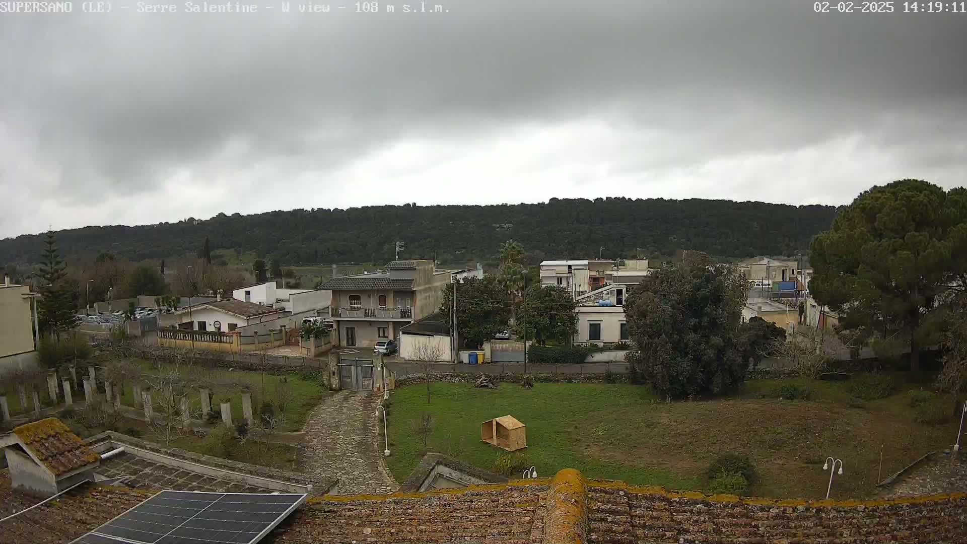 A view from a rooftop shows a small town nestled at the foot of a tree-covered hill under an overcast sky.