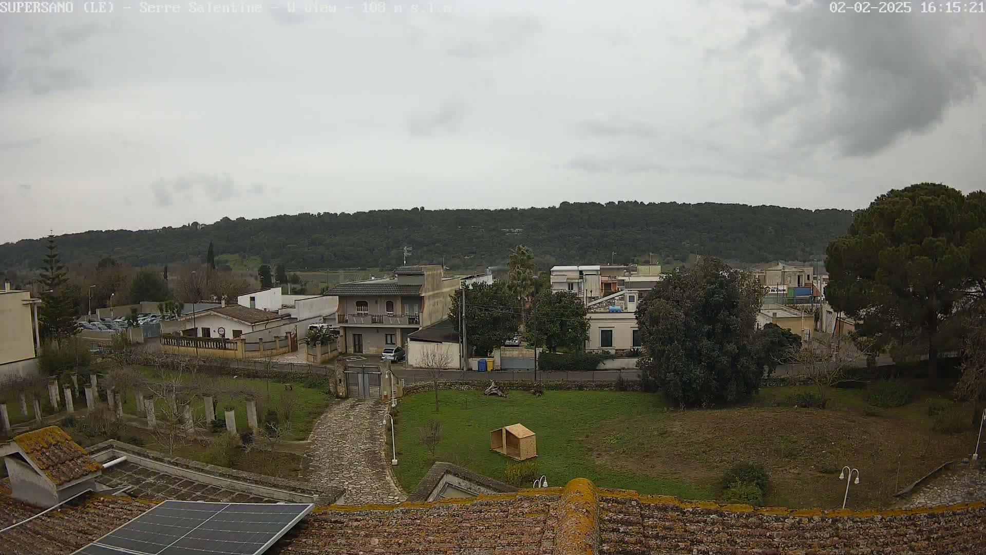 A cloudy day overlooks a residential area with low-lying buildings, a paved road, and a grassy yard with a small wooden structure, all situated in front of a wooded hill.