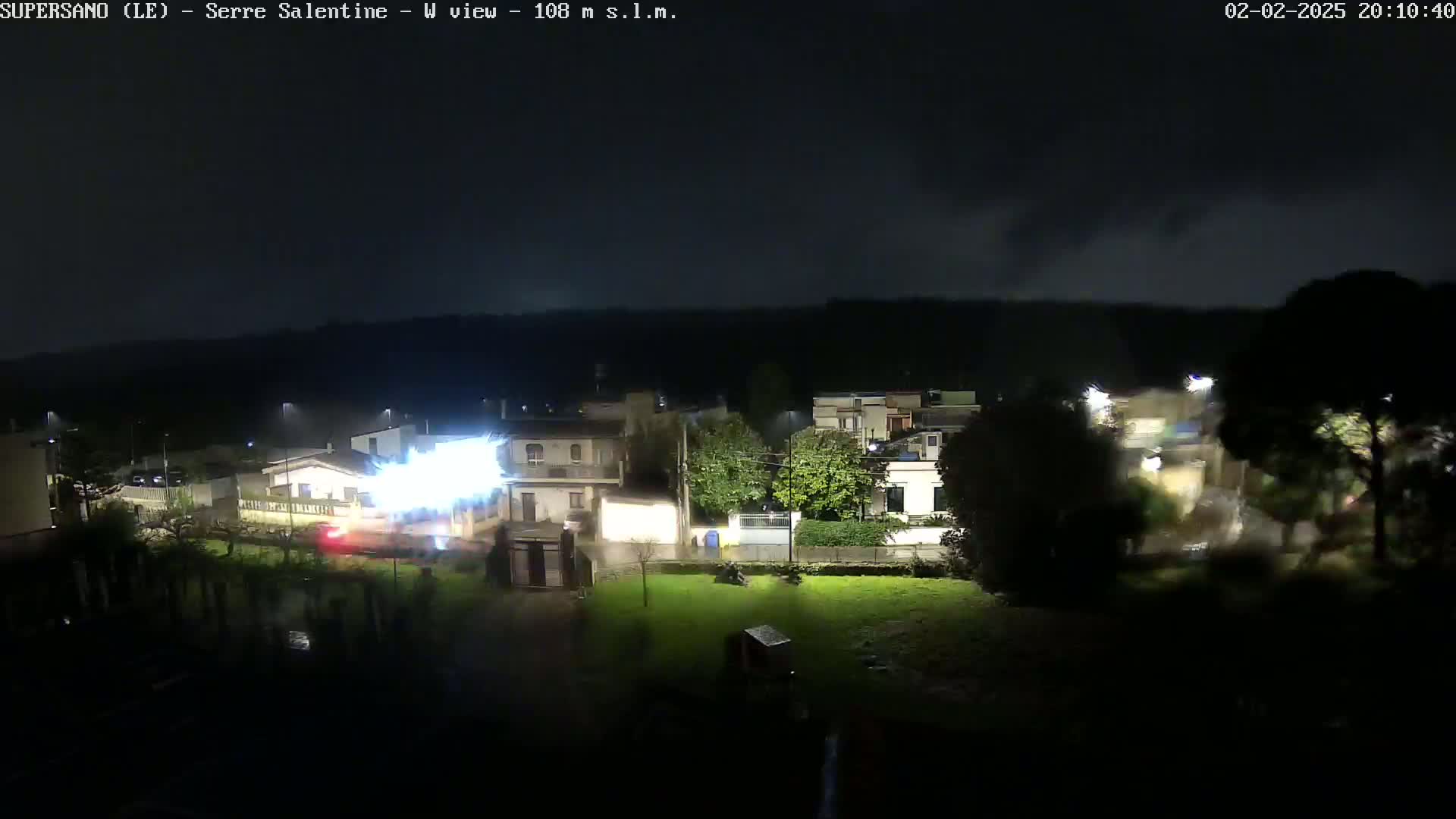 A nighttime view of a residential area with streetlights illuminated during a rainstorm, showing lightning striking near a street.