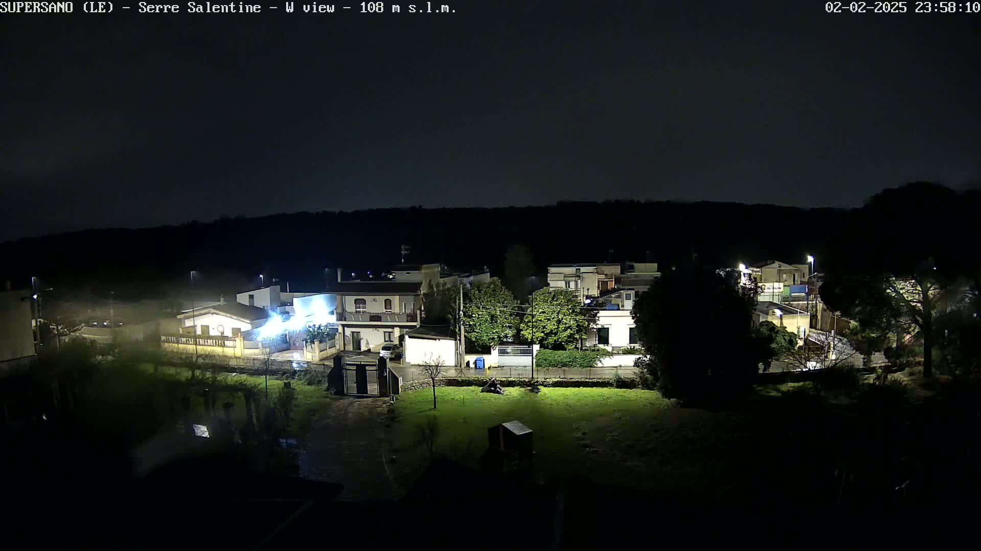 A nighttime, possibly rainy, overhead view of a residential area with houses and streetlights illuminated against a dark sky.