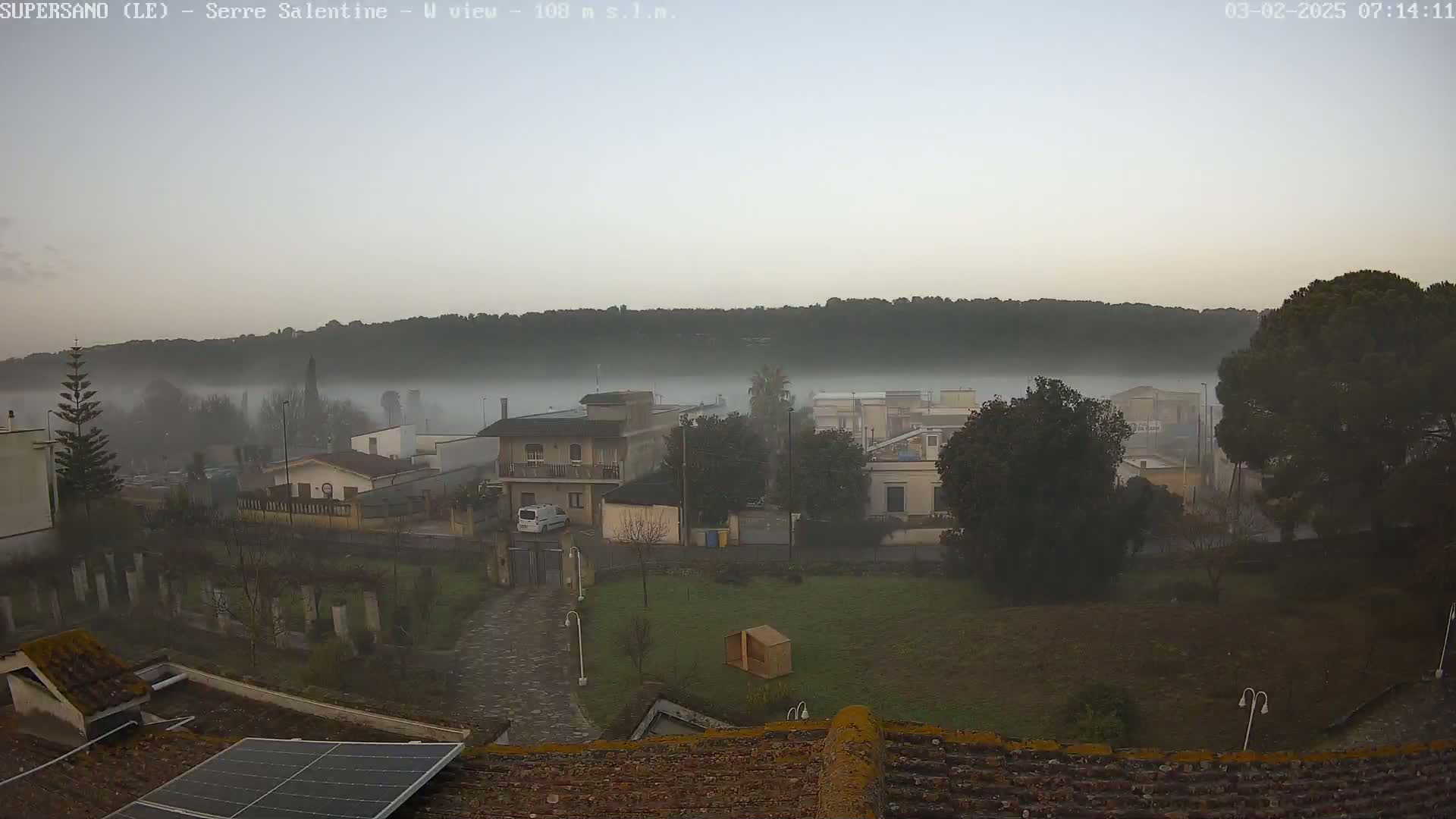 A foggy morning scene shows a residential area with low-lying buildings and trees, overlooking a hilly landscape shrouded in mist.