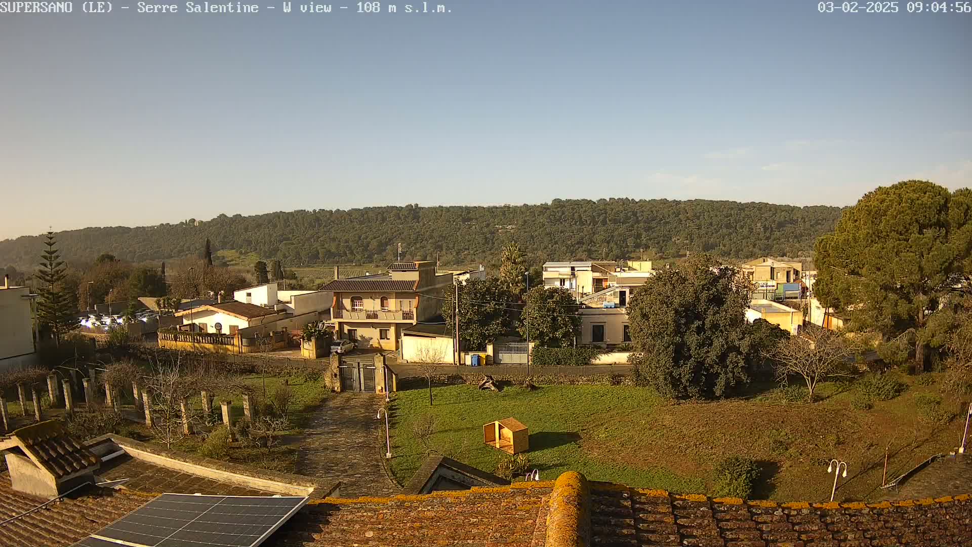 A clear, sunny day reveals a small town nestled at the foot of a tree-covered hill, viewed from a rooftop with a solar panel.