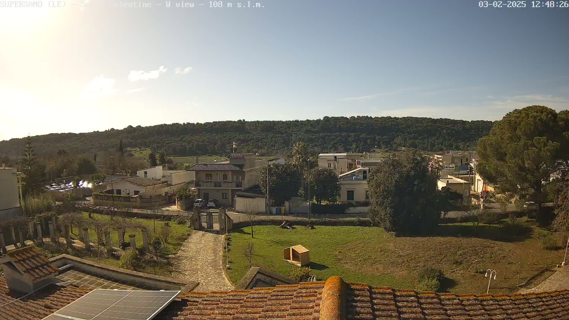 A sunny day reveals a small town nestled against a hillside, viewed from a rooftop with a solar panel.
