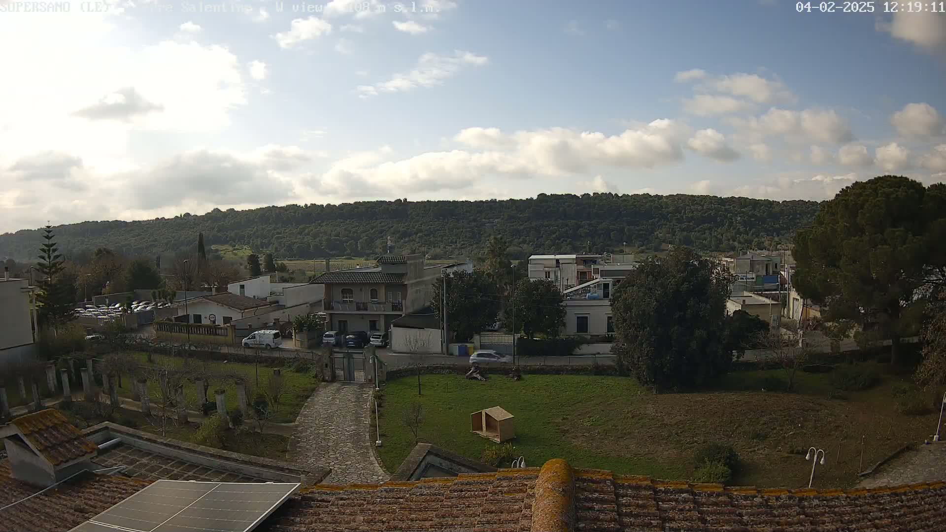 A partly cloudy sky overlooks a residential area nestled against a wooded hillside.