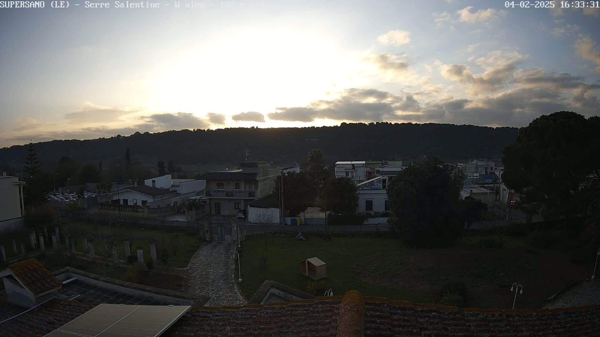A partly cloudy sunset illuminates a residential area nestled at the foot of a hill.