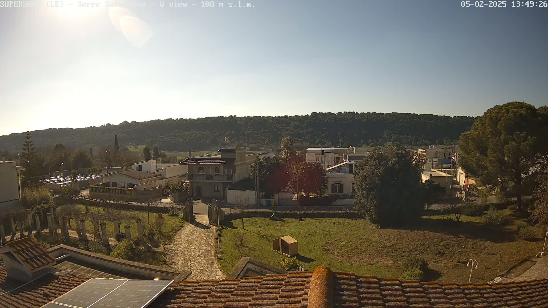 A sunny day overlooking a residential area nestled against a wooded hillside, viewed from a rooftop with a solar panel.