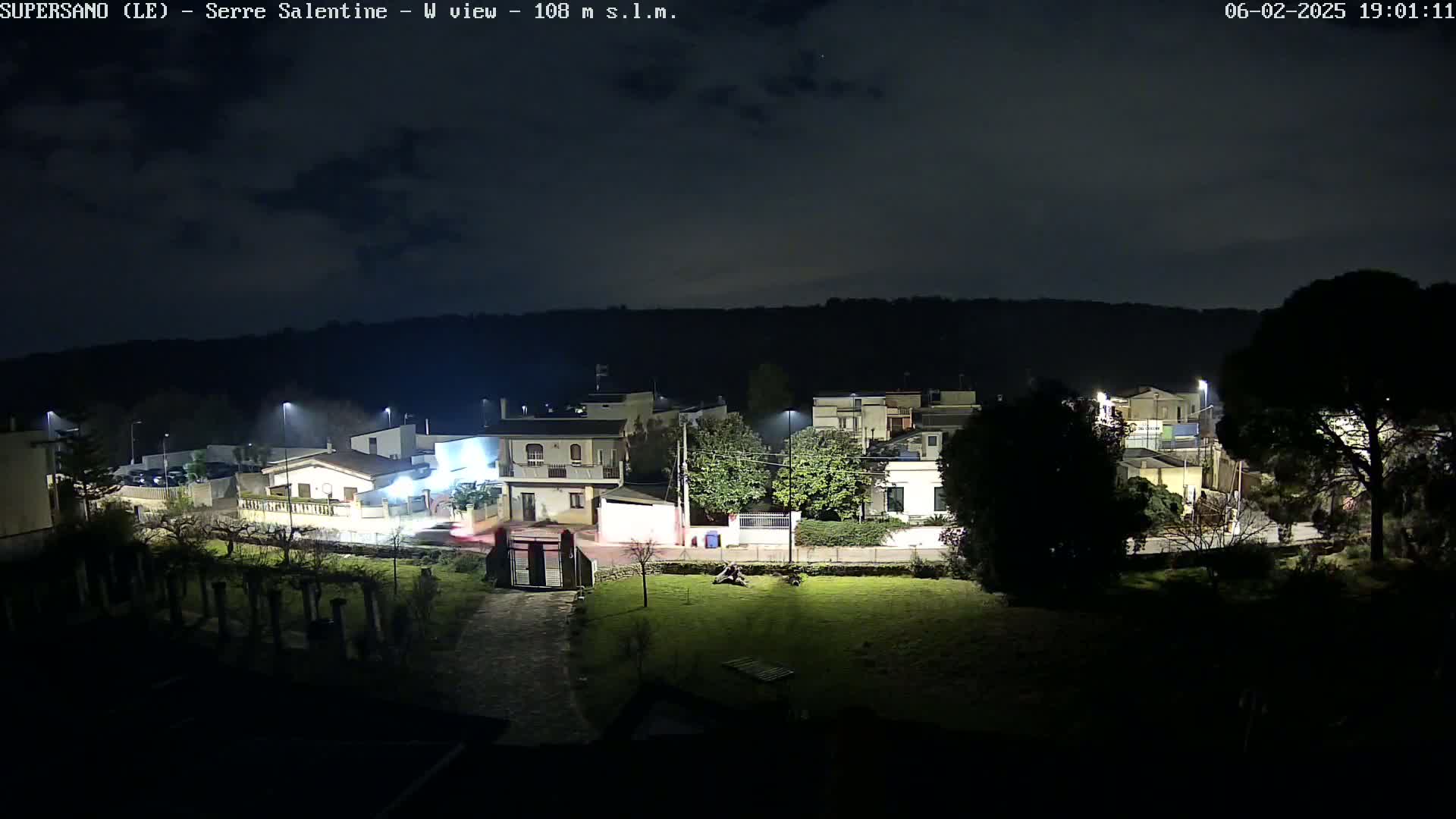 A nighttime view of a residential area with streetlights illuminating houses and a grassy yard, under a mostly clear sky.