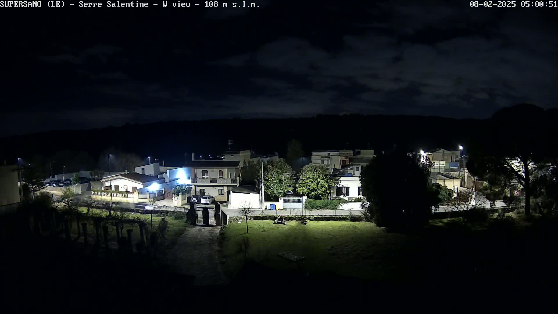 A nighttime, cloudy view of a residential area with illuminated houses and streetlights, partially obscured by trees.