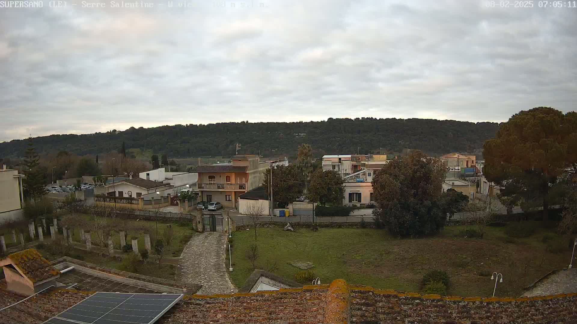 A mostly cloudy day reveals a residential area nestled against a backdrop of rolling hills, viewed from a rooftop with a solar panel.