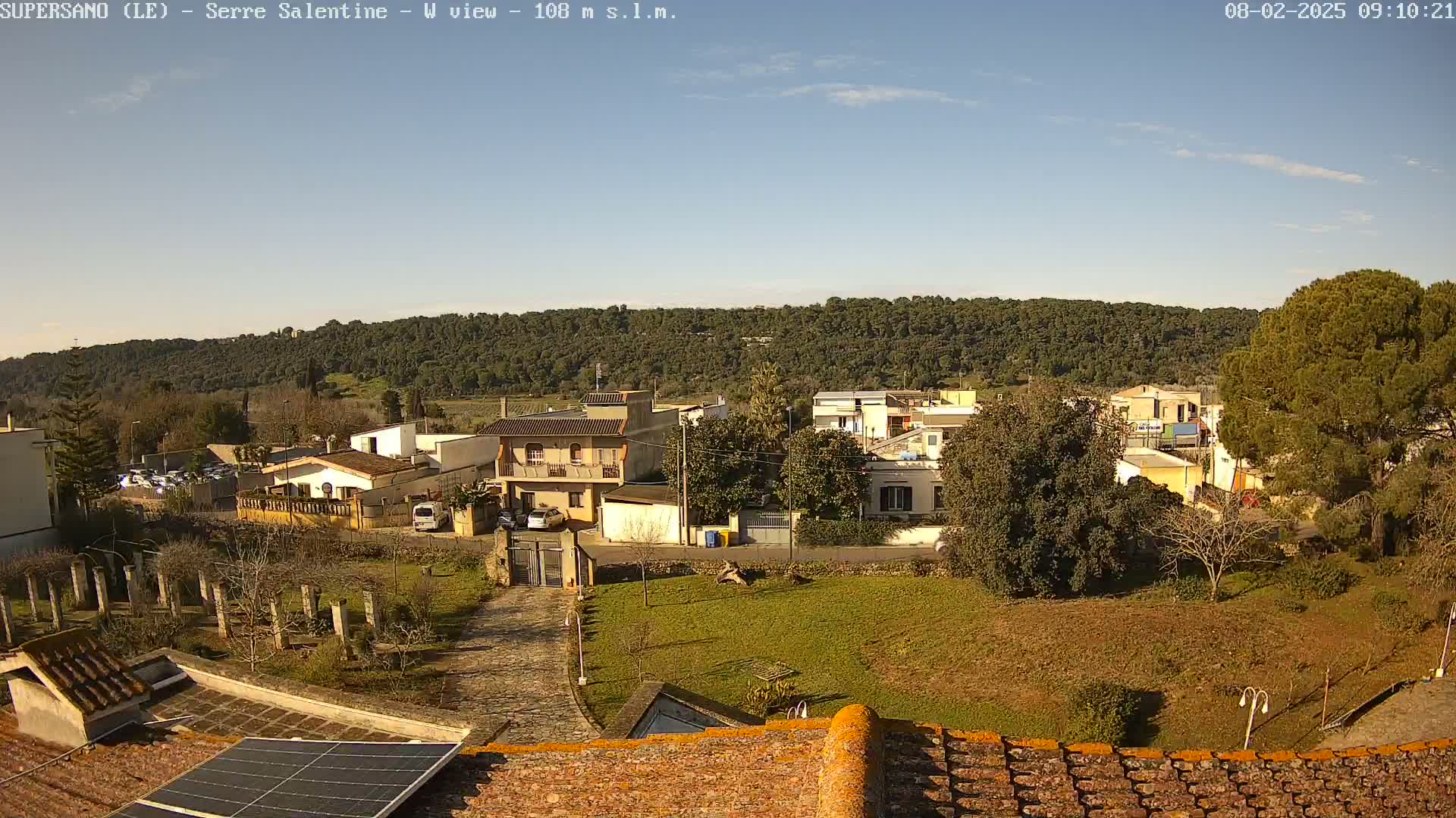 A sunny day reveals a village nestled below a hill covered in trees, viewed from a rooftop with a solar panel.