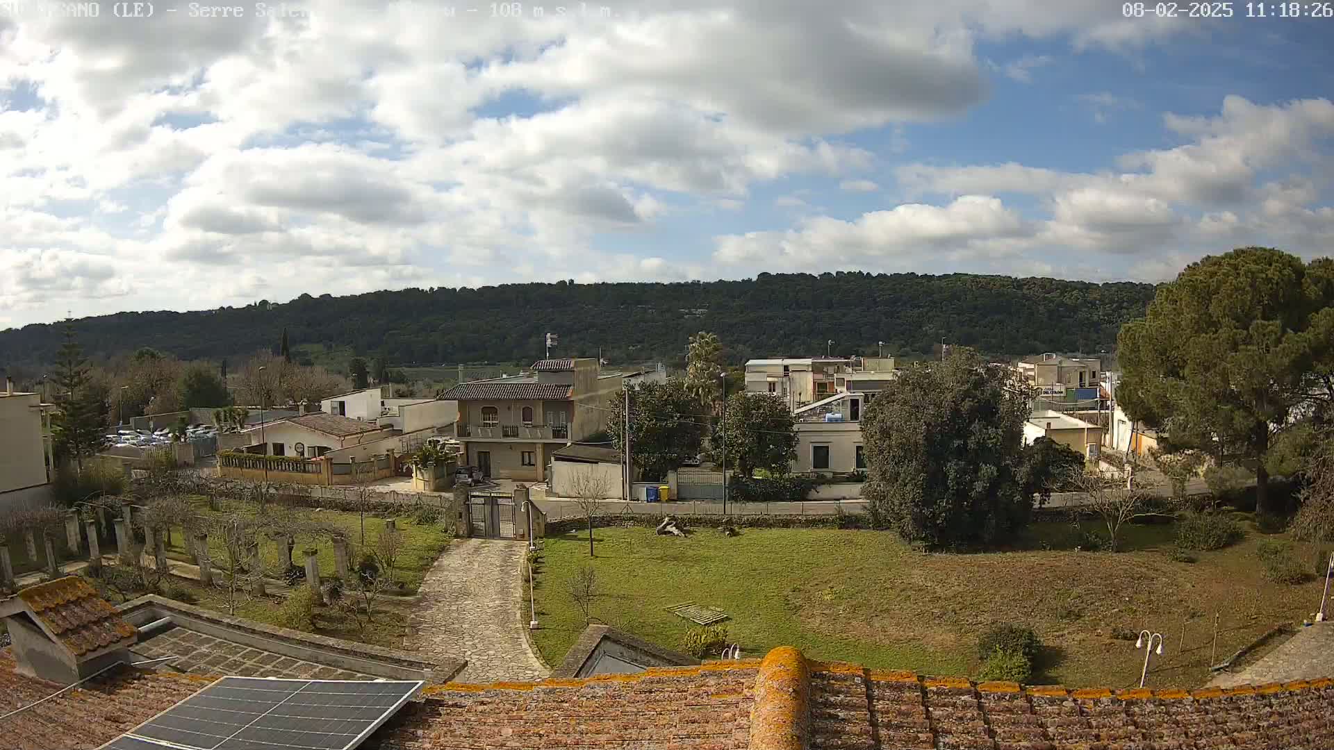 A partly cloudy day reveals a residential area nestled against a backdrop of verdant hills, viewed from a rooftop with a solar panel.