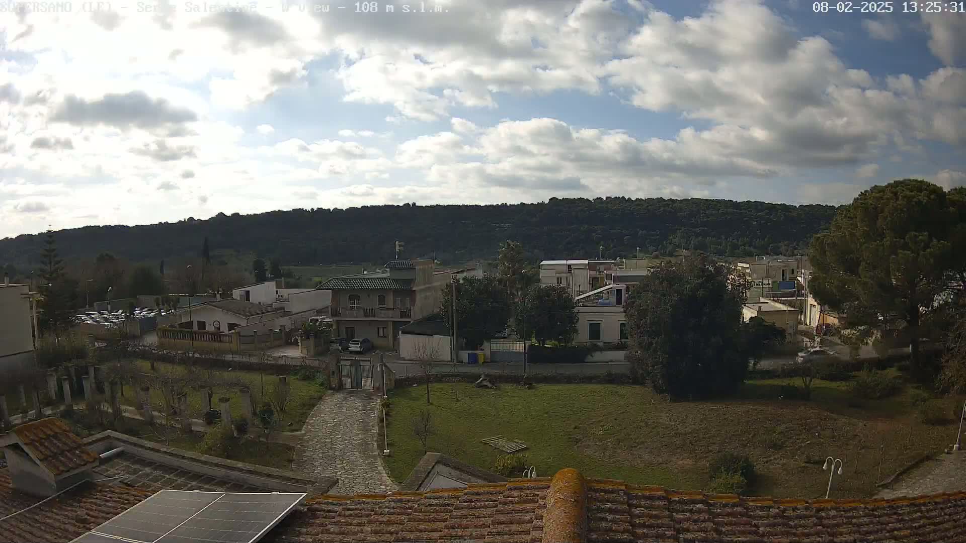 A partly cloudy day reveals a residential area nestled against a verdant hillside, viewed from a rooftop with a solar panel.