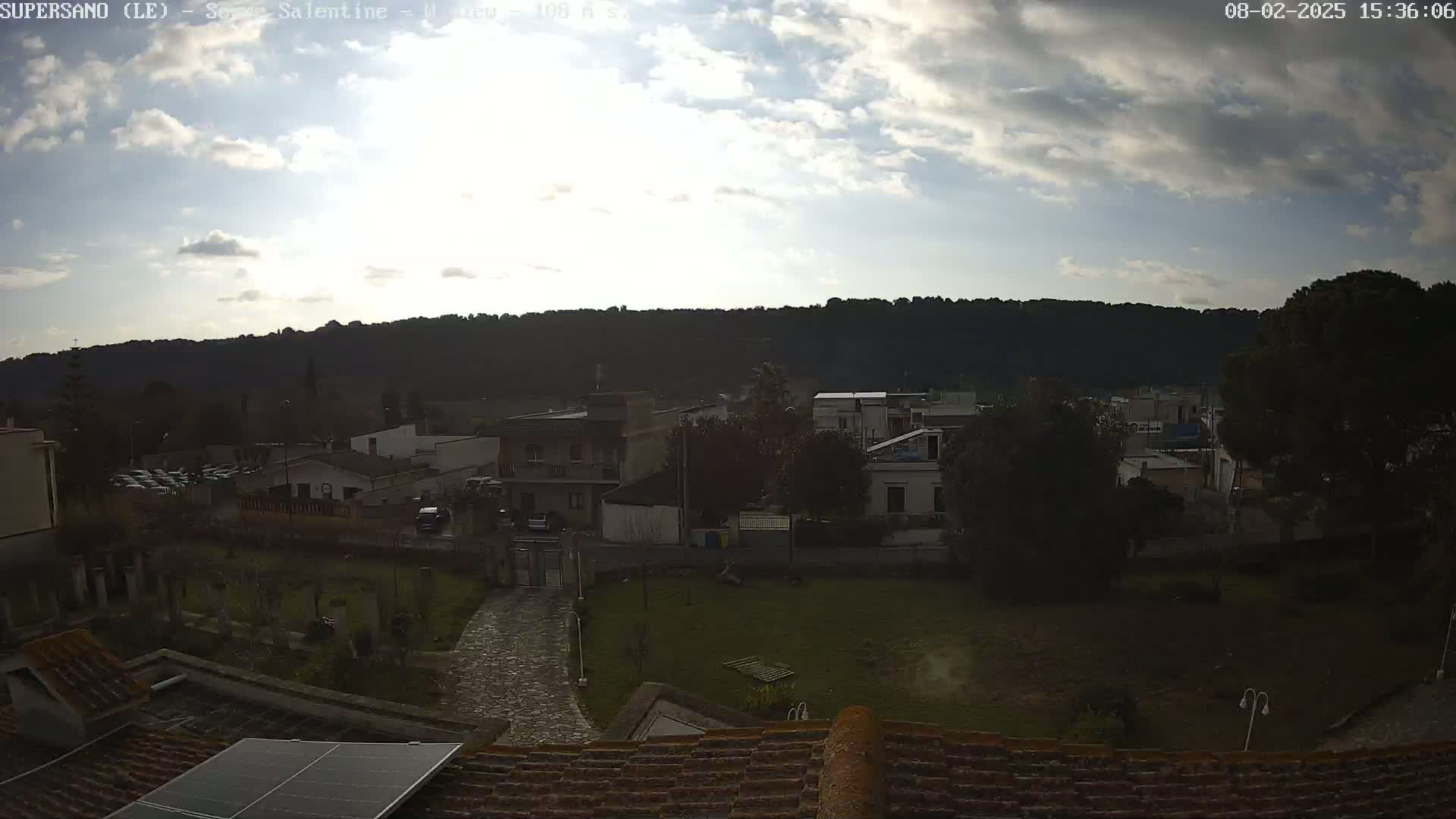 A partly cloudy sky overlooks a small town nestled against a dark, forested hillside.