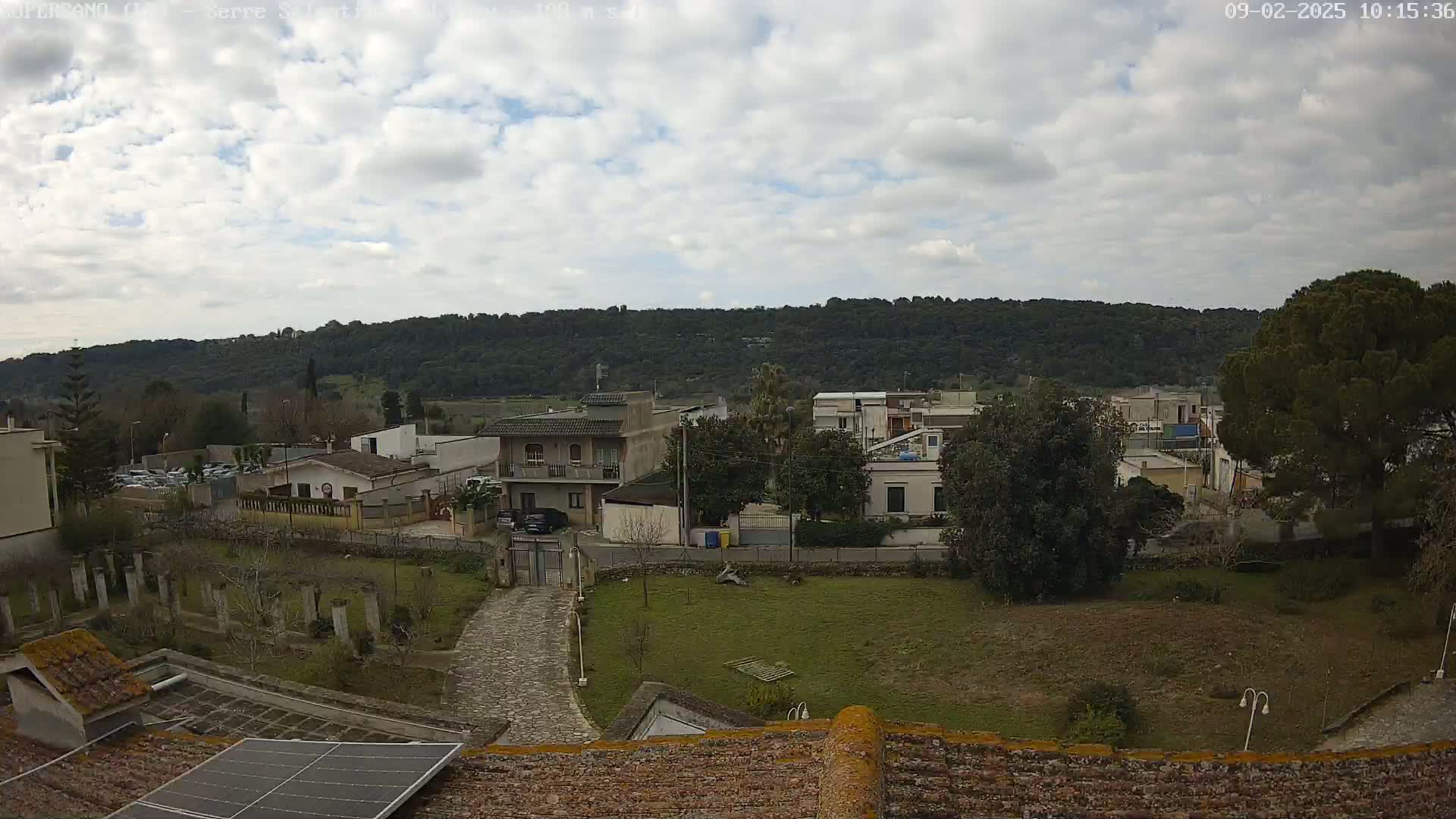 A partly cloudy day reveals a view of a residential area with low buildings nestled against a backdrop of a verdant hill.
