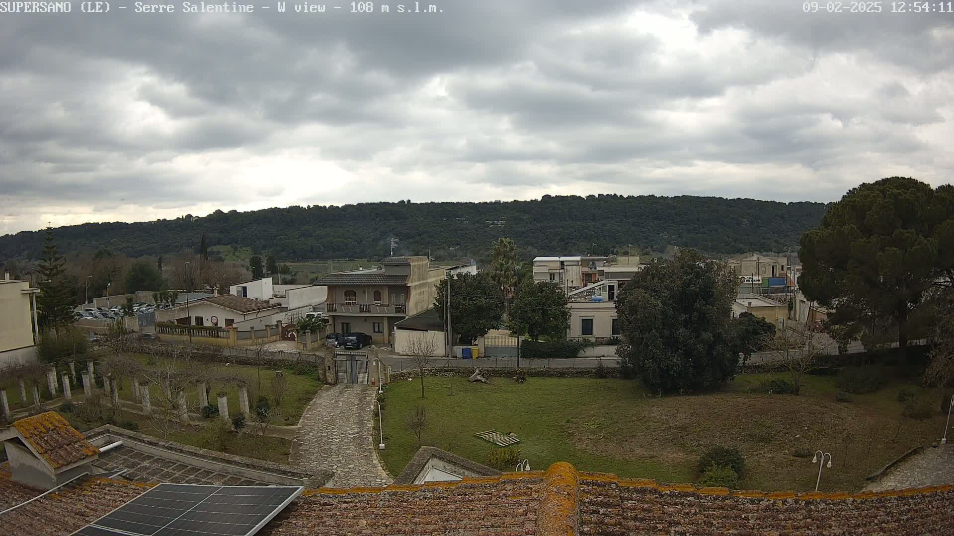 A view of a town nestled beneath a cloudy sky, showing residential buildings, a paved road, and a grassy area with a stone path leading to a gated entrance.