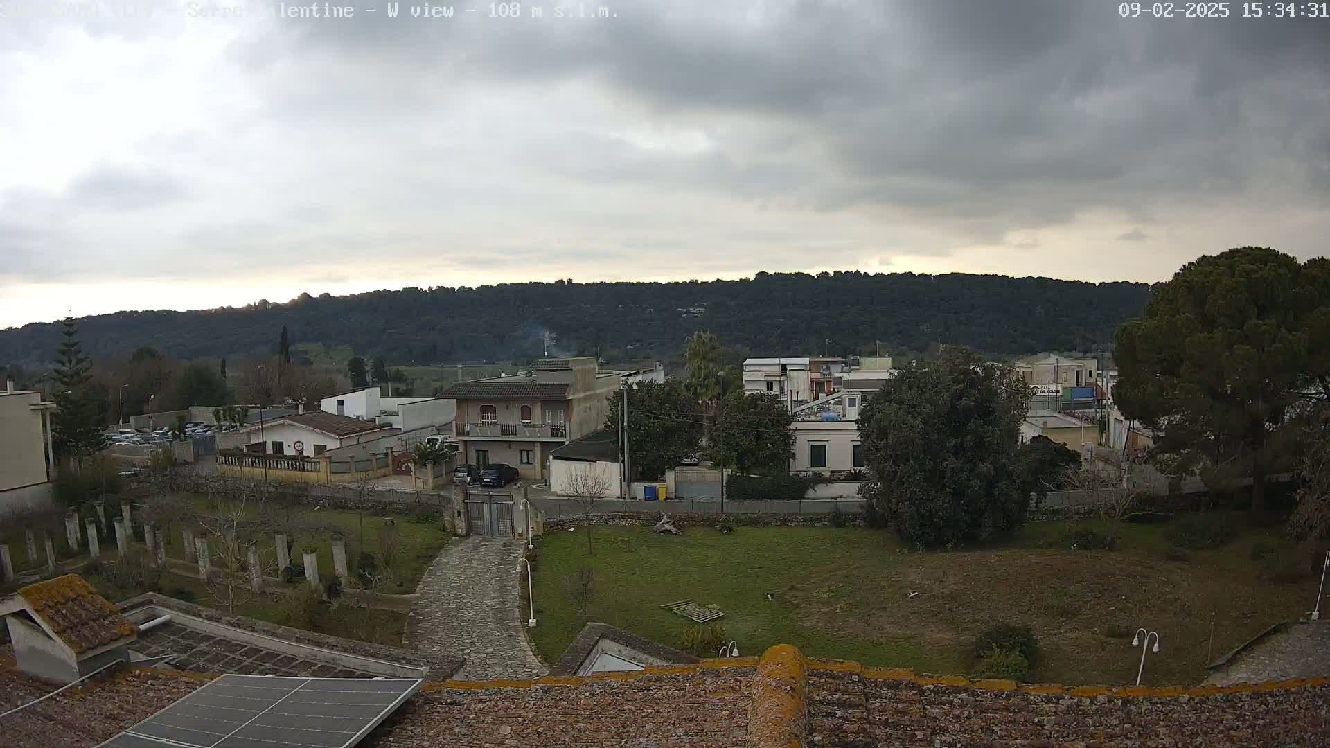 A view of a residential area with overcast skies, showing houses, trees, and a paved pathway leading to a gated entrance, all situated against a backdrop of distant hills.