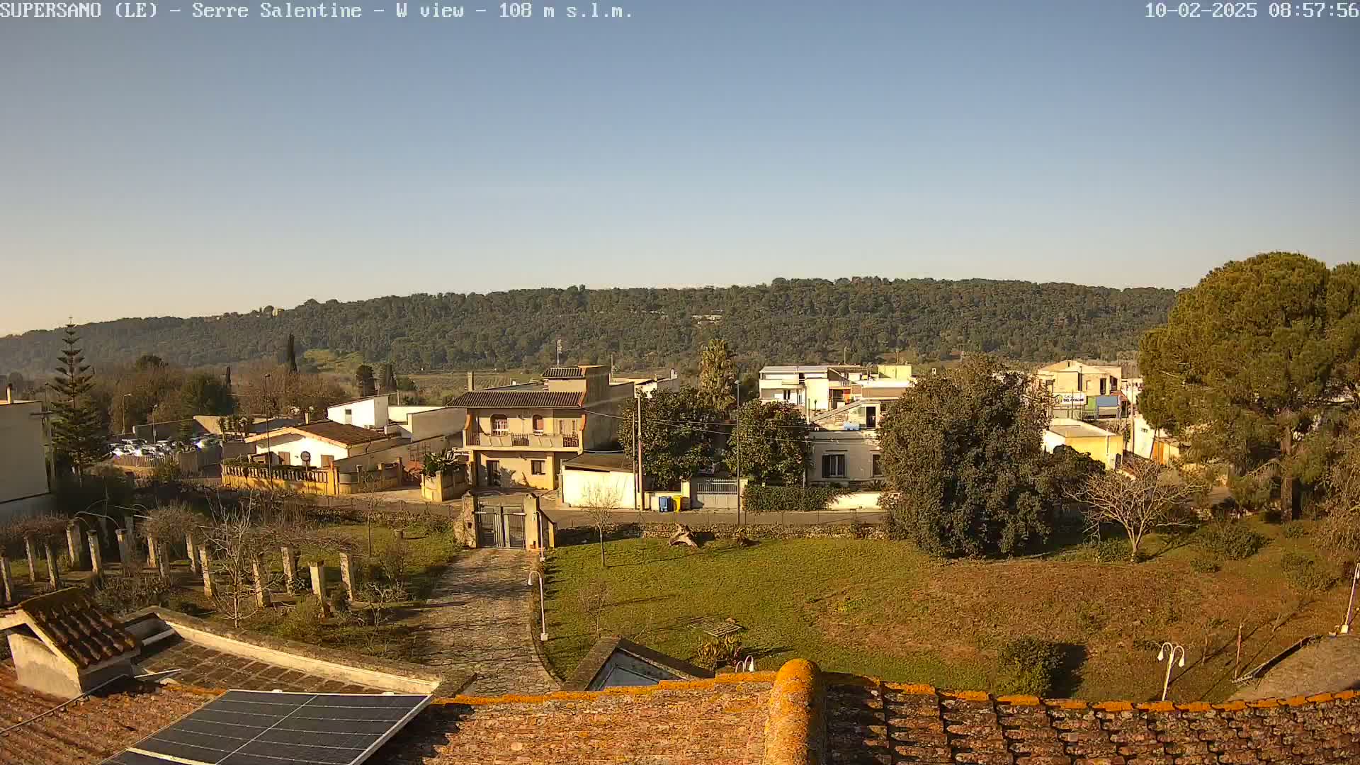 A sunlit view of a residential area nestled against a tree-covered hillside under a clear blue sky.