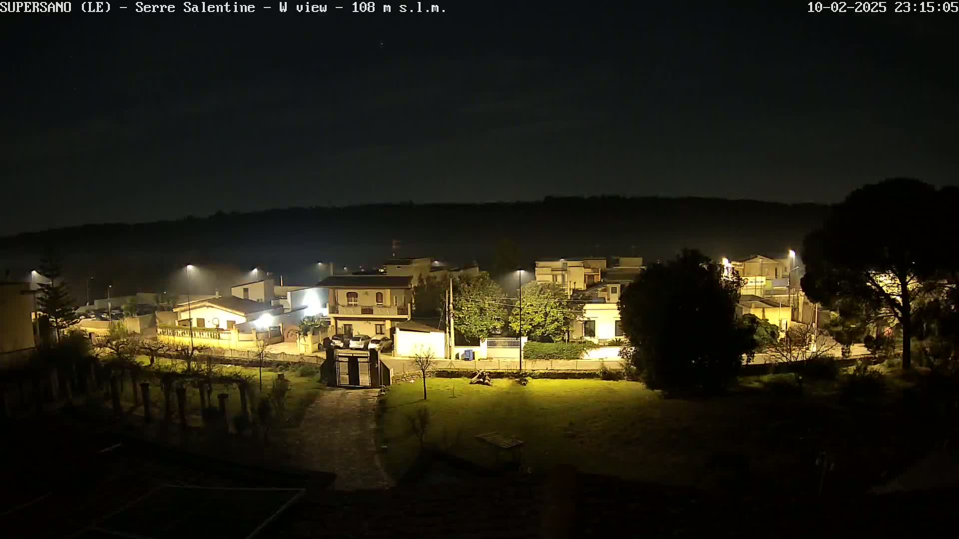 A nighttime view of a residential area illuminated by streetlights, with a hazy or foggy atmosphere.