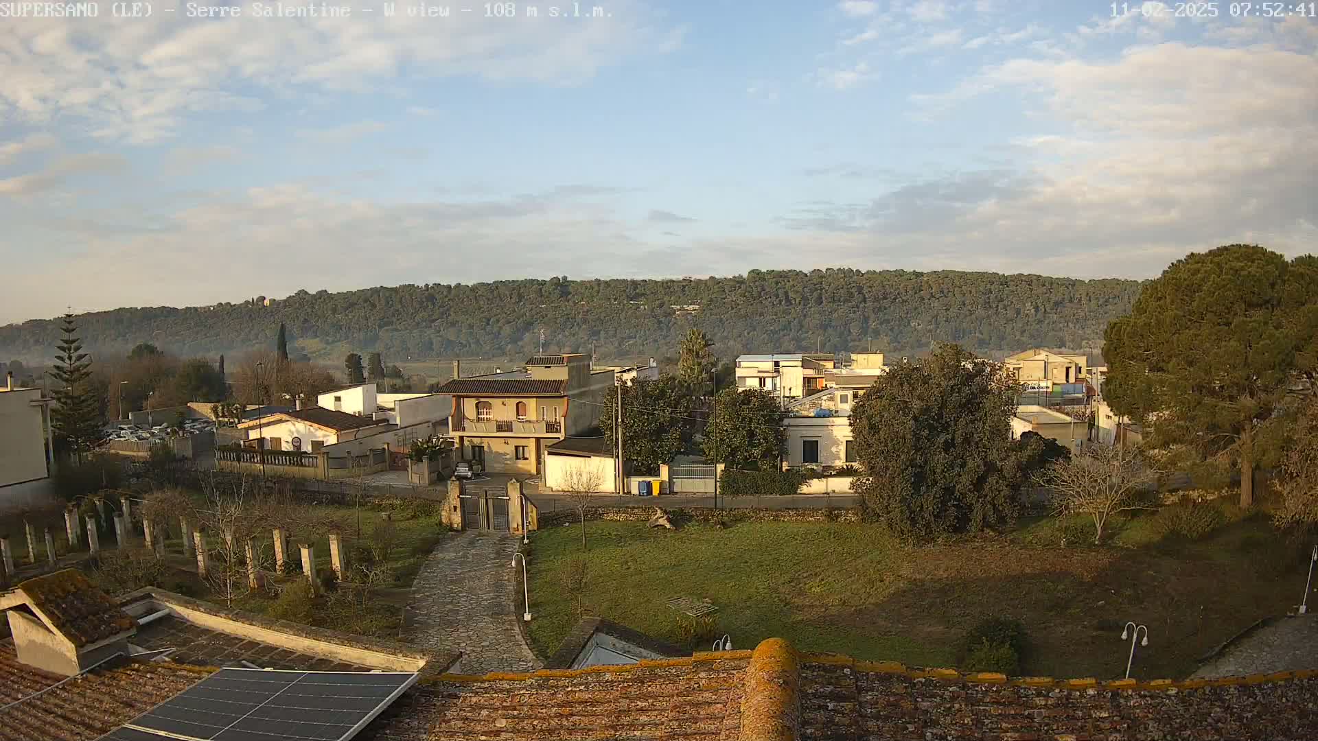 A partly cloudy morning reveals a small town nestled below a tree-covered hill, viewed from a rooftop with a solar panel.