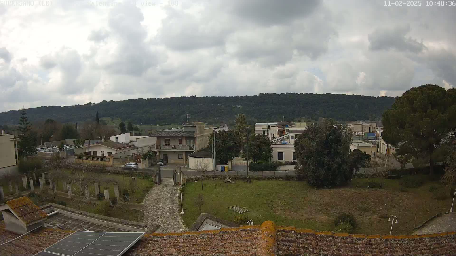 A cloudy day overlooks a residential area with a stone path leading to a green lawn, surrounded by buildings and trees, with a hill in the background.