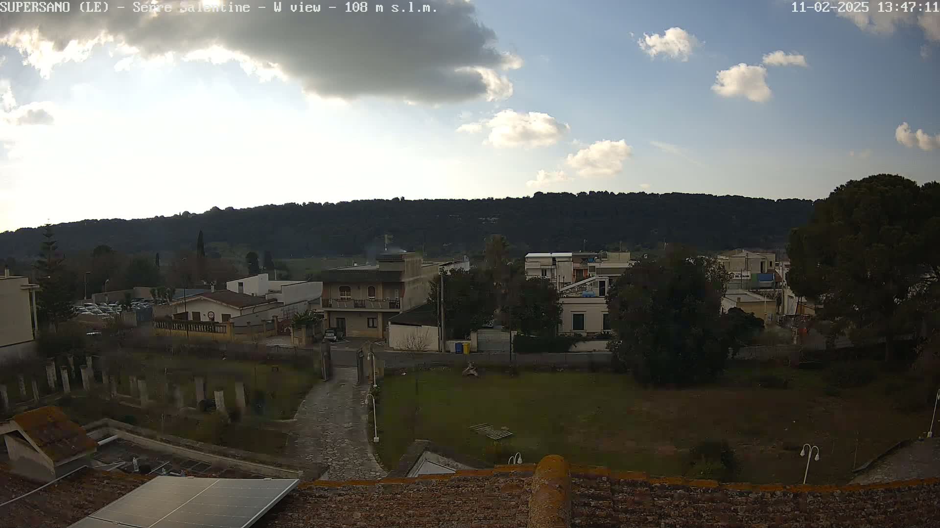 A partly cloudy day reveals a view of a town nestled below a tree-covered hill.