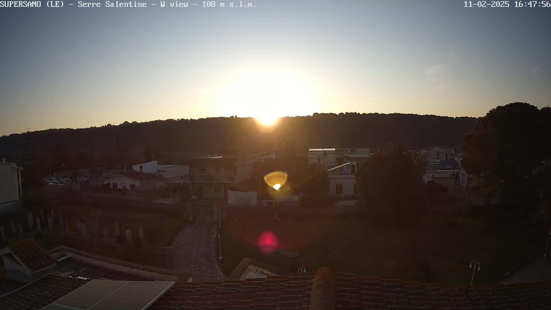 A bright sun sets over a small town nestled against a hillside, casting a golden glow on the buildings and landscape.