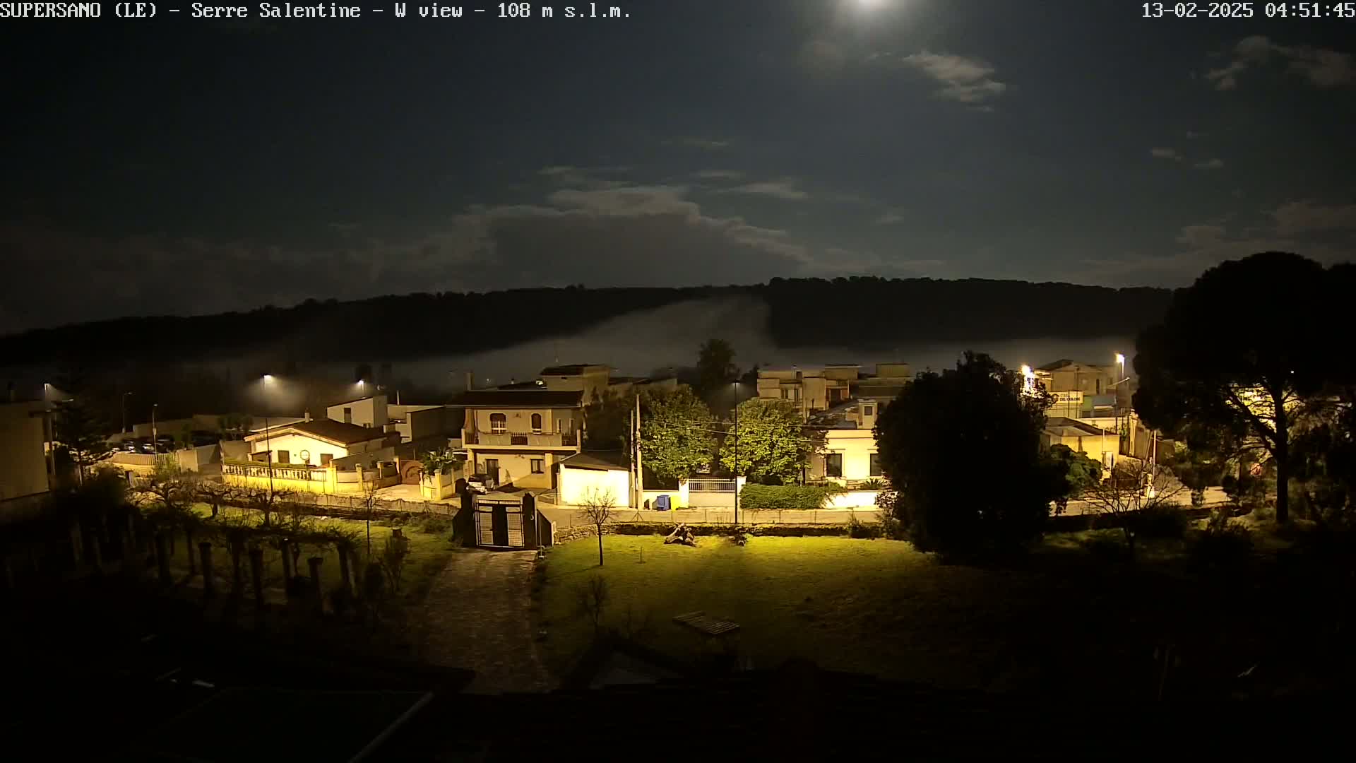A nighttime view of a town nestled in a valley, illuminated by streetlights, with low-lying fog partially obscuring the surrounding hills under a partly cloudy moonlit sky.