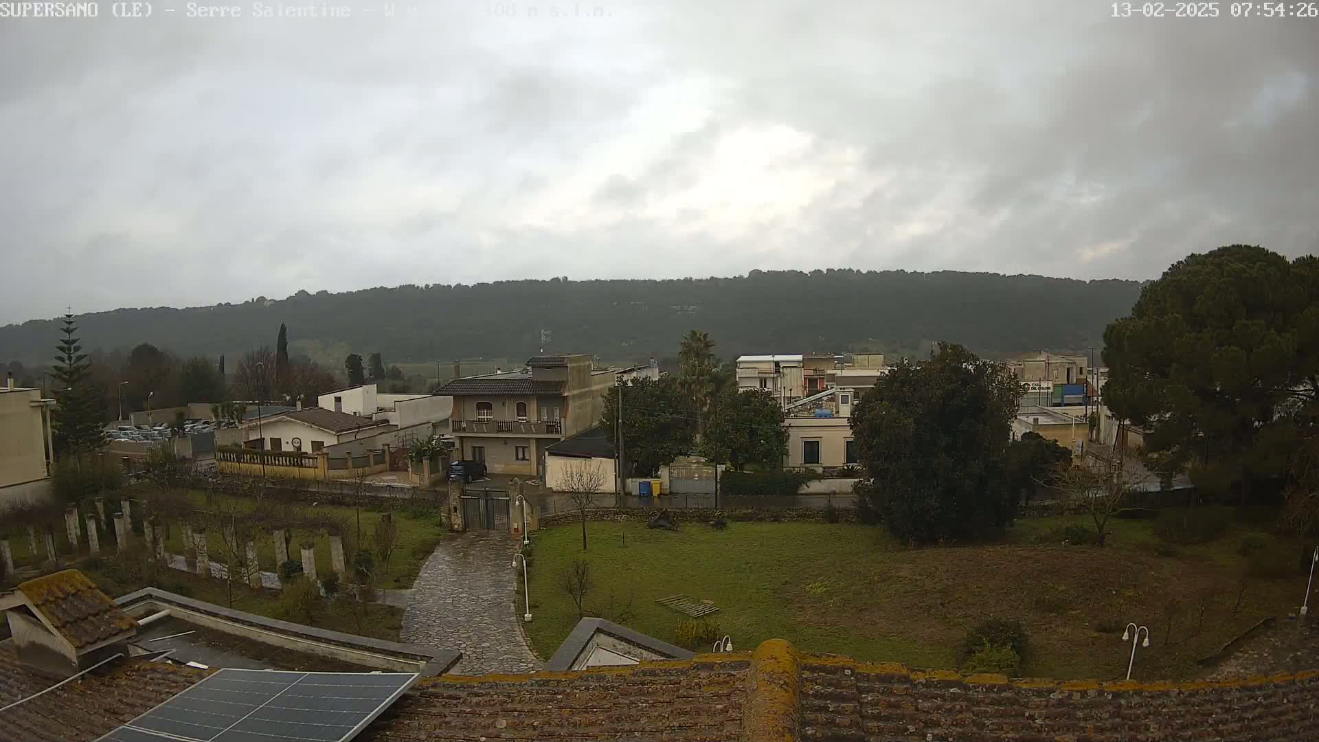 A view of a residential area with overcast skies shows houses, trees, and a paved path, with a hillside in the background under a gray sky.