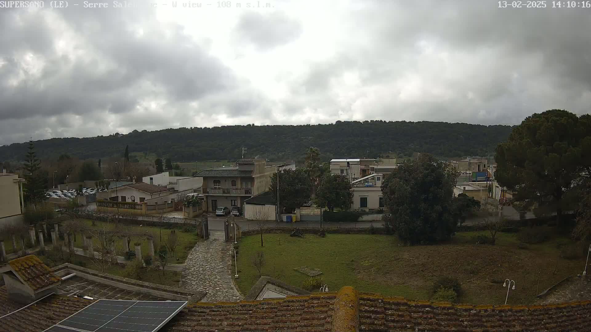 A cloudy day overlooks a residential area nestled beside a tree-covered hill, with a paved path and small garden visible in the foreground.