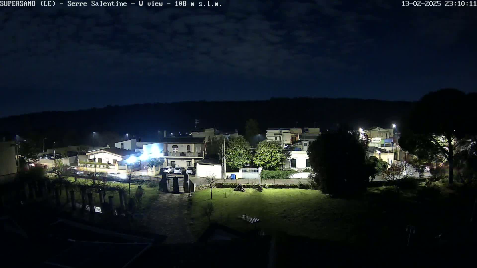 A nighttime view of a residential area under a partly cloudy sky shows houses and cars illuminated by streetlights.