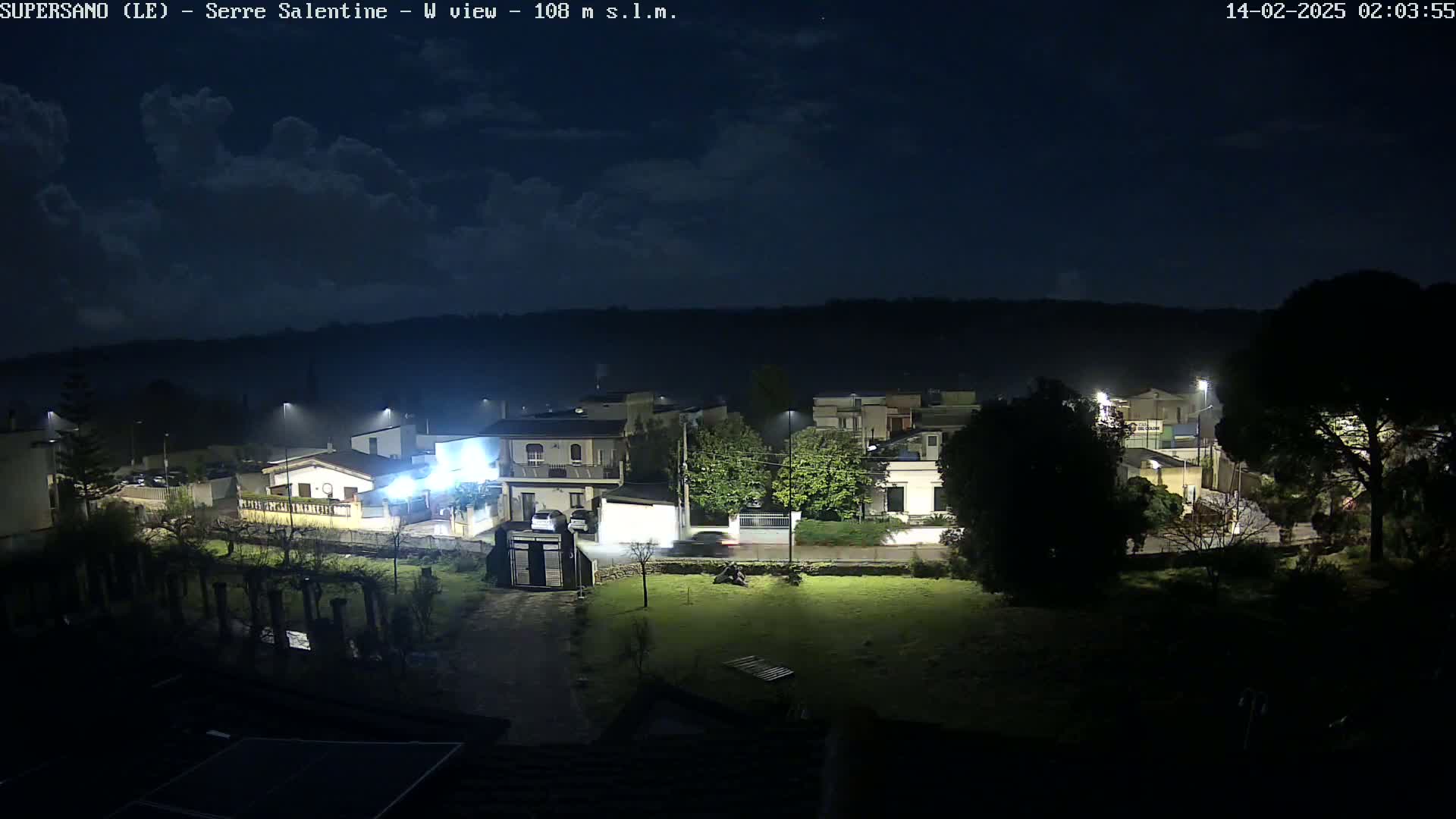 A nighttime view of a residential area under a partly cloudy sky, illuminated by streetlights.