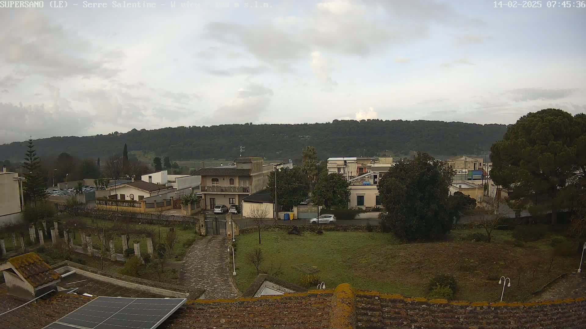 A view of a residential area with overcast skies, showing houses, trees, and a paved driveway leading to a gate, all nestled against a backdrop of rolling hills.