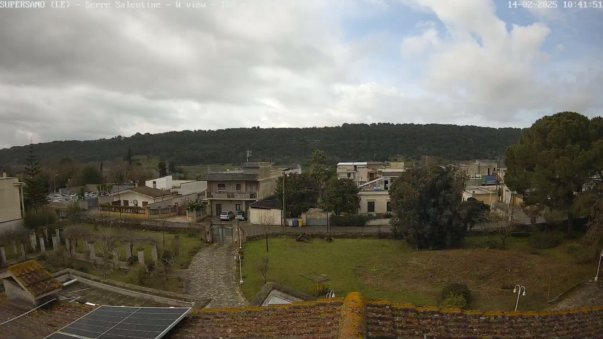 A partly cloudy day reveals a view of a residential area nestled against a tree-covered hillside, seen from a rooftop with a solar panel.