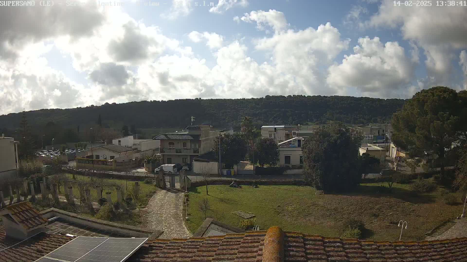 A partly sunny day reveals a view of a small town nestled against a wooded hillside, seen from a rooftop with a solar panel.
