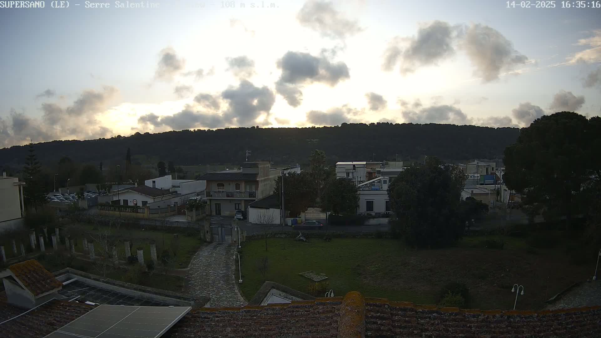 A cloudy sunset illuminates a residential area nestled against a hill, with a paved path leading to a gated entrance.