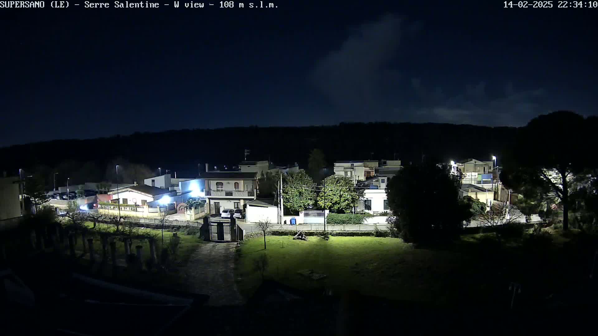 A nighttime, high-angle view of a residential area with houses illuminated by streetlights under a partly cloudy sky.