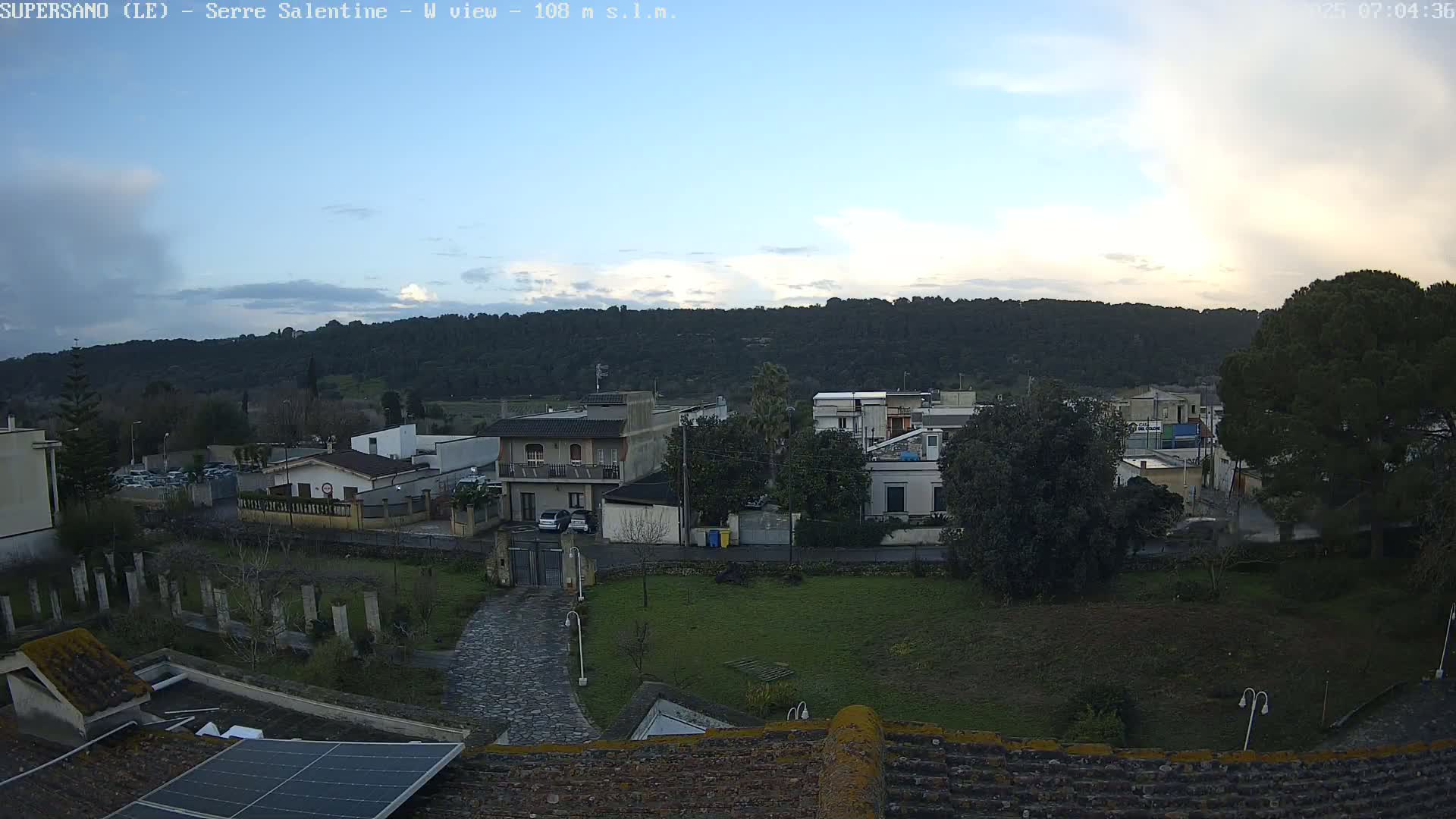 A mostly cloudy, late afternoon view of a residential area nestled against a tree-covered hill.