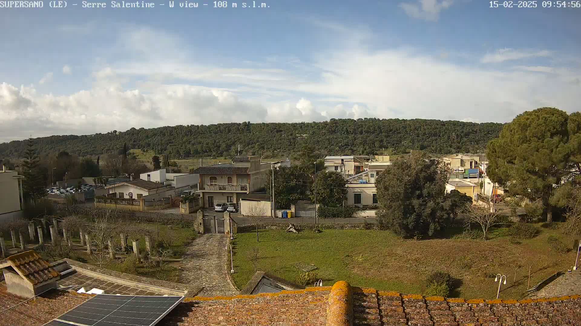 A partly cloudy day reveals a view of a small town nestled at the base of a wooded hill, seen from a rooftop with a solar panel.