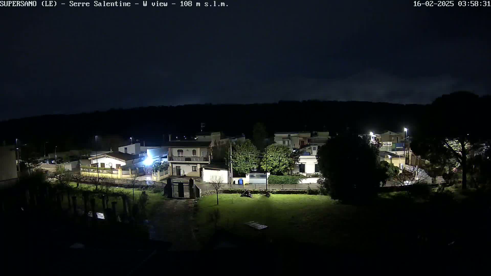 A nighttime view of a small town nestled against a dark hillside under a mostly clear, dark sky.