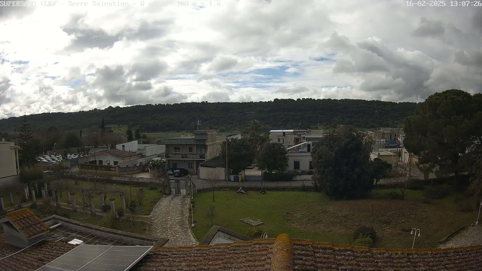 A cloudy day reveals a view of a residential area nestled at the base of a verdant hillside, seen from a rooftop with a solar panel.