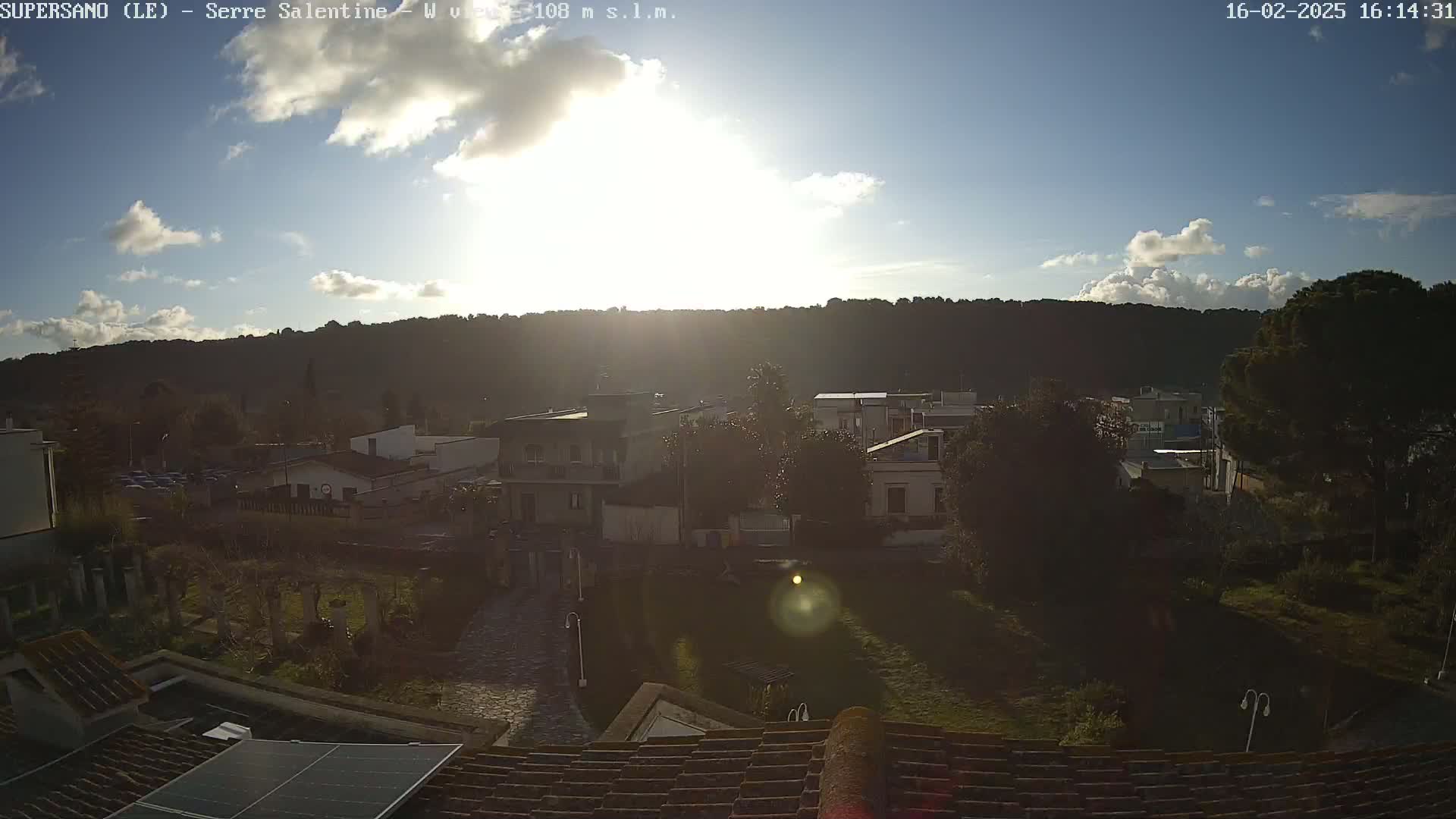 A partly sunny day reveals a residential area nestled below a hill, partially obscured by shadows and lens flare.