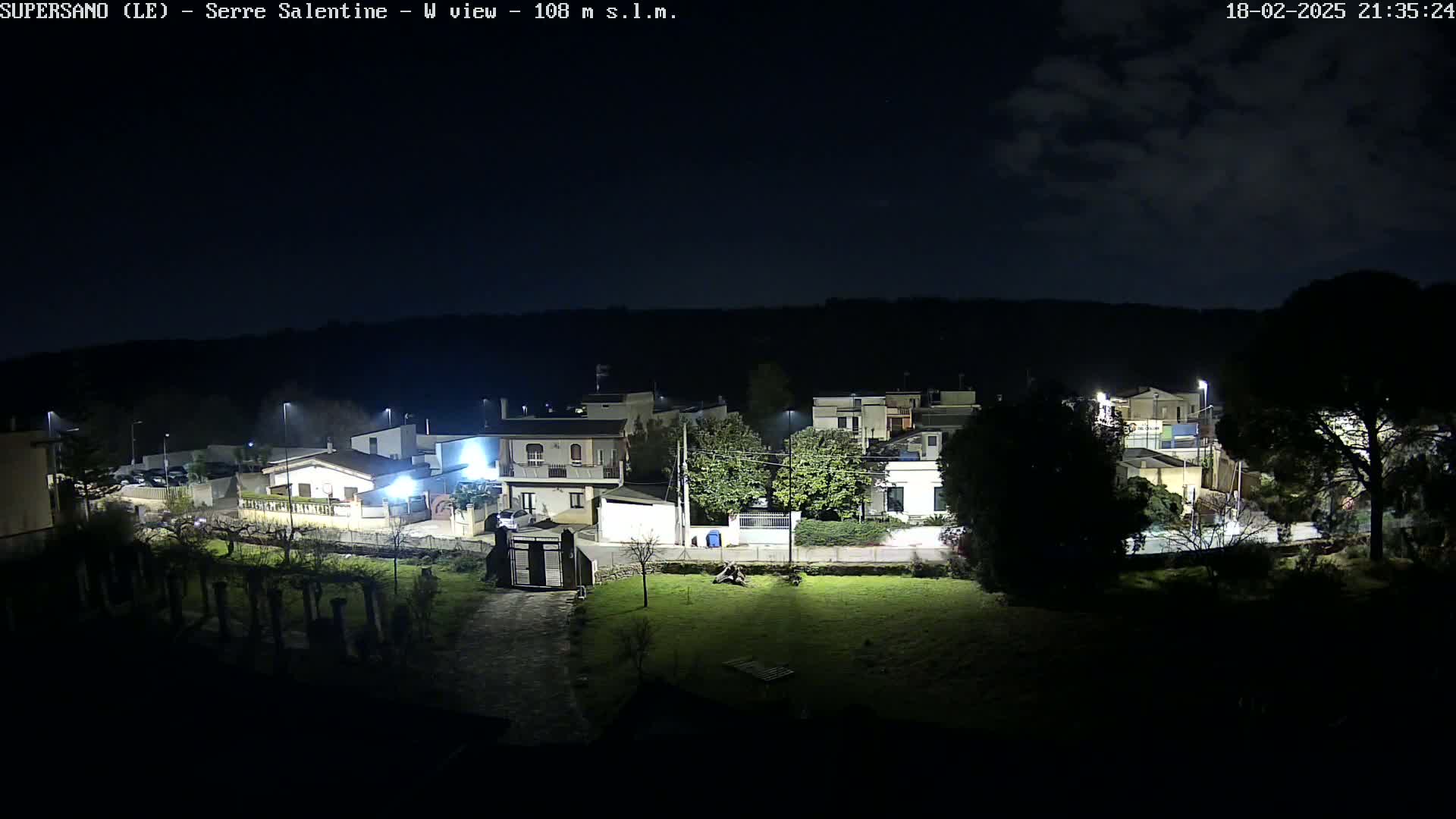 A nighttime view of a residential area under a partly cloudy sky, showing houses illuminated by streetlights and a dark, grassy area in the foreground.